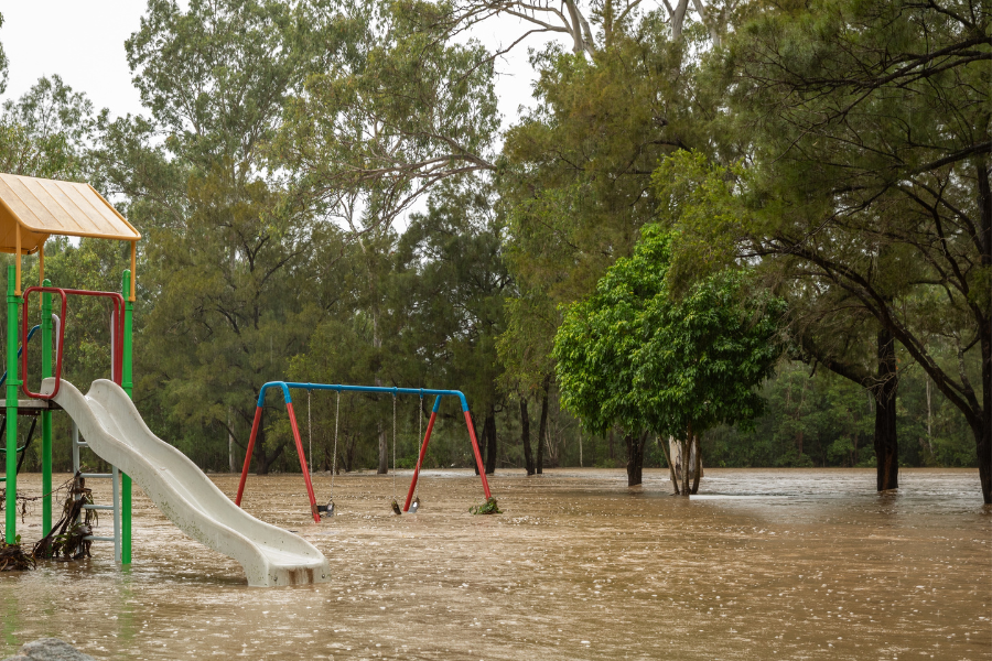 A playground with a slide and swings partially submerged in floodwaters, surrounded by trees.