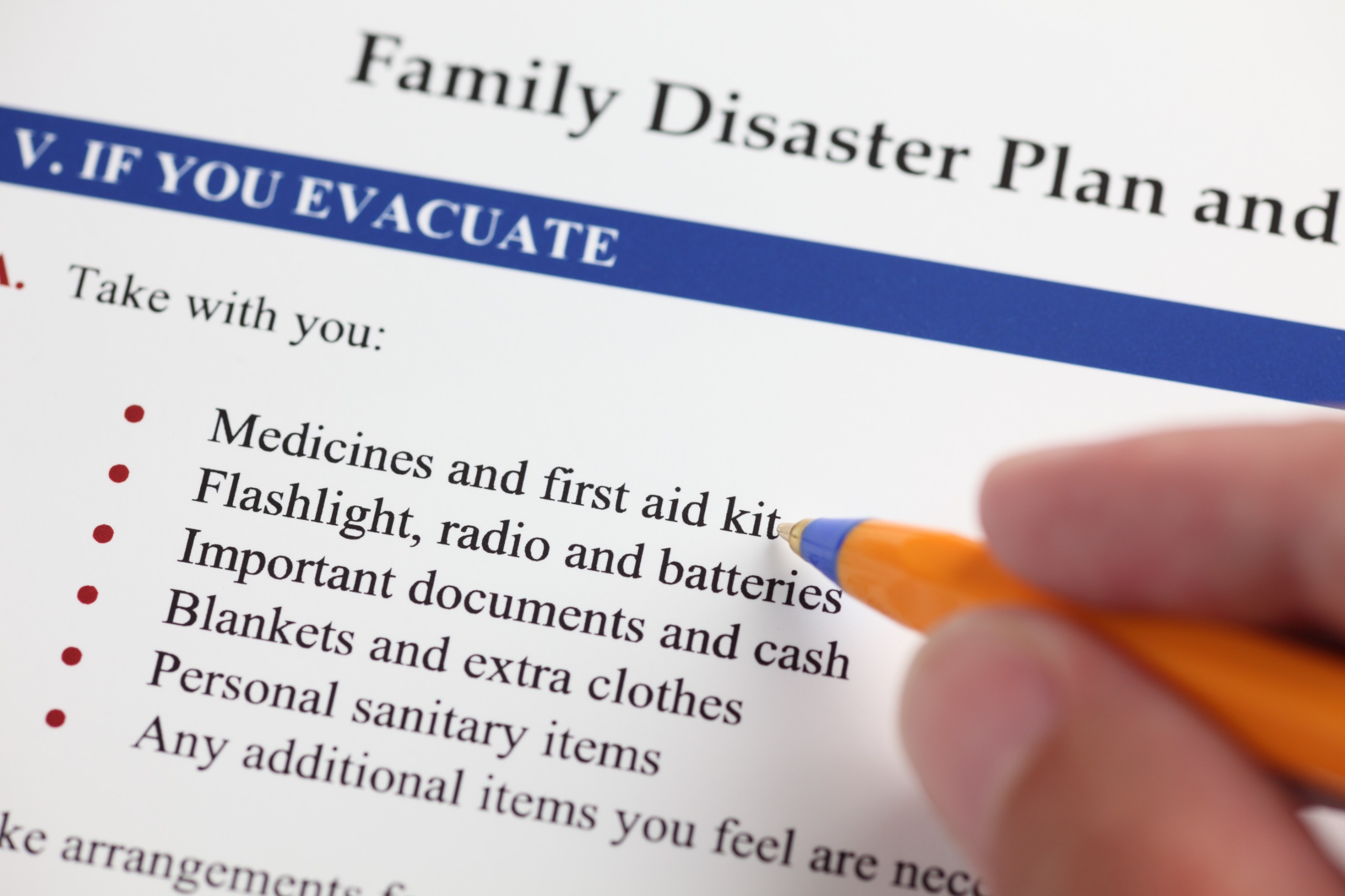 A hand holding a pen points to a list on a disaster plan document, highlighting essential items to take during an evacuation.
